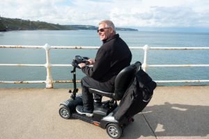 Middle aged male sits on his mobility scooter looking at the sea from the promenade at New Quay in Wales hoping to see dolphins or other wildlife, enjoying the freedom his scooter gives him to be able to get out and about unaided.